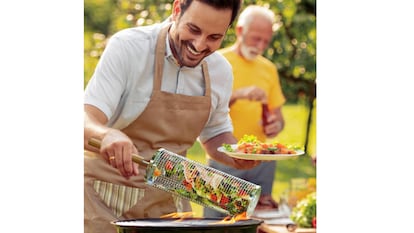 Un hombre cocina verduras a la parrilla con ayuda de una cesta de acero inoxidable.