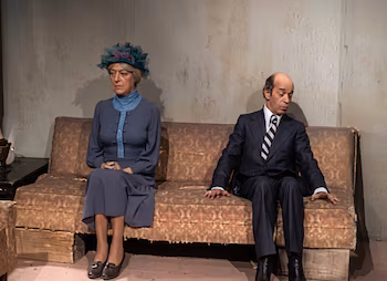 Un hombre calvo de traje y una mujer de vestido azul y sombrero se sientan distanciados en un sofá antiguo frente a una pared texturizada