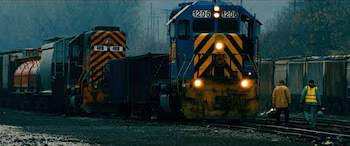 Dos trenes de carga con locomotoras azul y amarilla, y naranja y negra, en vías férreas con dos hombres caminando en primer plano